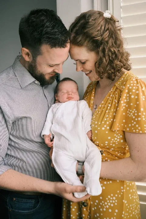 Smiling parents holding their sleeping newborn beside a window